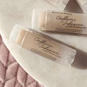 May include: Close-up of several clear tubes of Basswood Beauty Caffeine Infusion coffee infused eye balm. The tubes have white caps and labels with black script. The labels read "all natural" and "net wt. 0.15 oz (4g)".