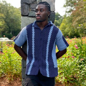 May include: A short-sleeved, button-down crochet shirt in shades of blue. The shirt has a collared neckline and a pattern of alternating dark and light blue vertical stripes. The garment is worn by a person standing outdoors.