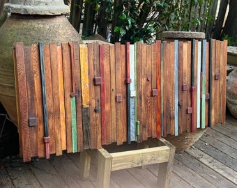 May include: A rectangular wooden art piece composed of vertical planks in various colors, including red, orange, green, and gray. Small, square wooden blocks are affixed to the planks. The artwork is displayed outdoors, resting on a wooden stand.