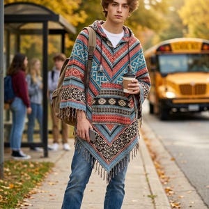 May include: A hooded poncho featuring a vibrant, multi-colored geometric pattern in shades of red, blue, and black. The garment includes a front kangaroo pocket, a drawstring hood, and a fringed hemline, paired with blue denim jeans and sneakers.