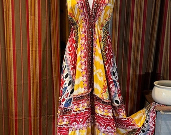 May include: A halter-neck maxi dress with a vibrant, colorful print. The dress features a deep V-neckline and a flowing skirt. The fabric is adorned with patterns in red, yellow, blue, and white. The dress is displayed on a mannequin.