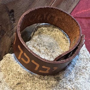 以下が含まれることがあります： 明るい茶色のヘブライ文字が入った茶色の革製カフブレスレット。ブレスレットは灰色の石の表面に置かれています。革は使い古されたように見え、素朴な外観になっています。