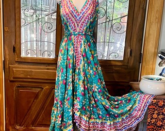 May include: A teal blue maxi dress with a floral print and a multi-colored trim. The dress has a V-neckline and a halter strap. The dress is on a white stand.