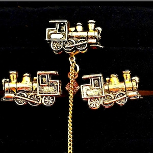 May include: A gold-colored train-themed jewelry set, including a pendant and matching earrings. The pendant hangs from a gold chain, and the earrings are designed as miniature train locomotives. The set is displayed against a dark background.