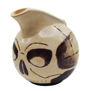 May include: A beige ceramic pitcher with a skull design. The pitcher has a rounded body with a spout and handle. The skull design is painted in dark brown, with cracks in the ceramic adding to the design.