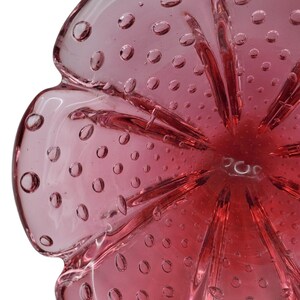 May include: A pink glass bowl with a flower-like shape. The bowl has a textured surface with small, evenly spaced bubbles throughout. The center of the bowl is a deeper shade of pink, with petal-like ridges extending outwards.