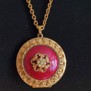 May include: A gold-coloured locket necklace featuring a red centre and a decorative floral design. The locket has a textured red centre with a gold filigree design and small pearl accents. The chain is a delicate gold link.