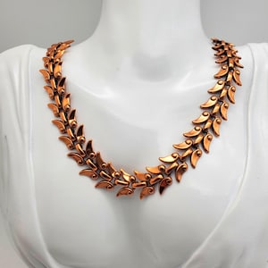 May include: A copper-toned necklace with a leaf-like design. The necklace is displayed on a white mannequin. The leaves are arranged in a repeating pattern, creating a symmetrical and elegant look. The metal has a warm, reflective sheen.