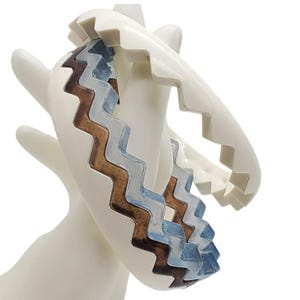May include: A white, zig-zag patterned bracelet with brown and blue accents. The bracelet is held by a white hand-shaped display. The bracelet's design features a layered effect with a unique, geometric pattern.