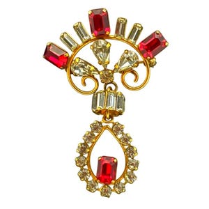May include: A vintage gold-toned brooch with a fan-shaped top adorned with red and clear rectangular and teardrop-shaped rhinestones. A teardrop-shaped pendant with a red rectangular rhinestone hangs below, surrounded by clear rhinestones. The brooch has a decorative, ornate design.