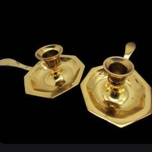 May include: Two vintage brass candle holders. Each holder has a hexagonal base with a handle and a cup for the candle. The candle holders are a shiny gold color and are set against a black background.