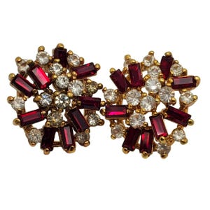 May include: A pair of gold-toned clip-on earrings. Each earring features a circular design with a cluster of clear and ruby-red rectangular and round-cut rhinestones. The earrings are set against a white background.