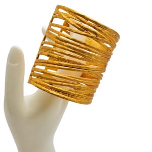 May include: A wide cuff bracelet crafted from gold-toned metal, featuring a series of horizontal, textured wire-like bands arranged in an open, cage-like structure that wraps around the wrist area with a consistent, repeating pattern.