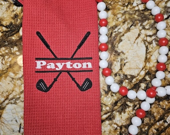 May include: Red golf towel with black trim and a white embroidered design of two crossed golf clubs and the name "Payton". The towel is hanging from a white and red beaded necklace with a red tassel.