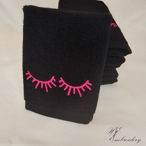 May include: Black face cloths with embroidered pink eyelashes. The cloths are stacked, with the front one showing the full design. The embroidery is a bright pink colour, contrasting with the dark fabric. The overall theme is beauty and self-care.