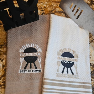Puede incluir: Dos toallas de cocina con diseños bordados. Las toallas son marrones y blancas con rayas marrones. El bordado dice "Dad's Barbecue Best in Town" con una parrilla y utensilios.