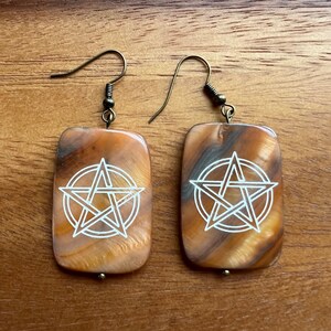 Puede incluir: Un par de pendientes con colgantes rectangulares veteados en naranja y marrón. Cada colgante presenta un diseño de pentagrama blanco. Los pendientes tienen ganchos para las orejas de color bronce y pequeñas cuentas doradas.