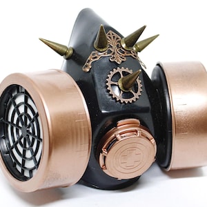 Könnte beinhalten: Eine schwarze Steampunk-Gasmaske mit kupferfarbenen Filtern und goldfarbenen Akzenten. Die Maske hat mehrere Stacheln und Zahnräder.