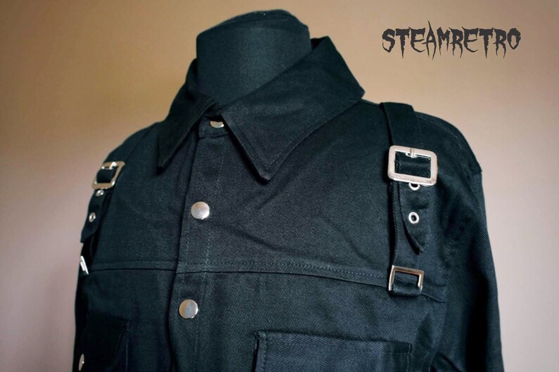 May include: A black long-sleeved shirt with a button-down collar and silver buckles on the shoulders. The shirt has a pocket on the left chest and a seam across the chest.