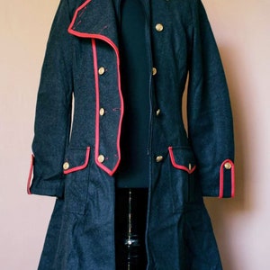 May include: A long, dark gray coat with a red trim and gold buttons. The coat has a stand-up collar and a zipper closure. The coat is on a mannequin.