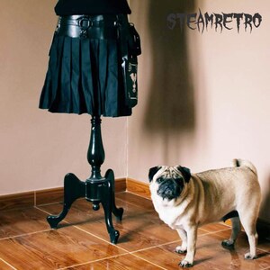 May include: A black skirt with pleats and a leather belt is displayed on a black stand. A small, light brown pug dog is standing on a wood floor.