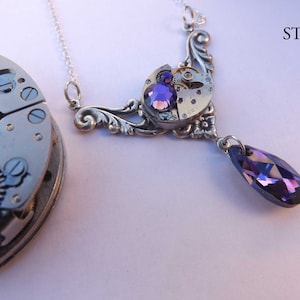May include: A silver necklace with a pendant featuring a vintage watch face and purple crystals. The pendant is attached to a silver chain with a teardrop-shaped purple crystal hanging from the bottom.