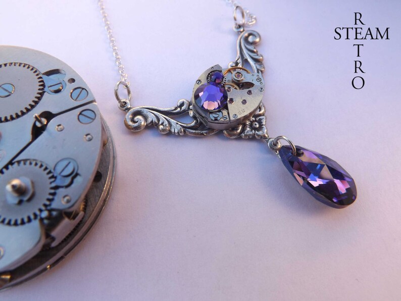 May include: A silver necklace with a pendant featuring a vintage watch face and purple crystals. The pendant is attached to a silver chain with a teardrop-shaped purple crystal hanging from the bottom.