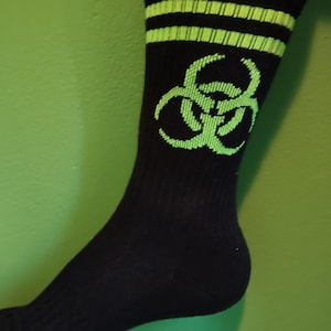 May include: A black crew sock featuring a bright lime green biohazard symbol centred on the side. The top of the sock includes two horizontal lime green stripes encircling the cuff, providing a distinct contrast against the solid black fabric.