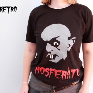 May include: A black t-shirt featuring a white and grey graphic of Count Orlok from the film Nosferatu. The text "NOSFERATU" is printed in red below the graphic.