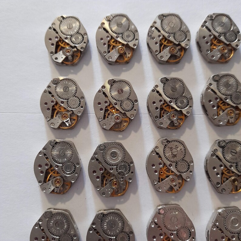 May include: A collection of 18 silver-toned watch movements with intricate gears and mechanisms. The movements are arranged in rows on a white surface.