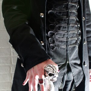 May include: A person wearing a black coat with a skull-shaped walking stick. The coat has a decorative design of black braid on the front. The person is wearing a ring with a red stone on their finger.