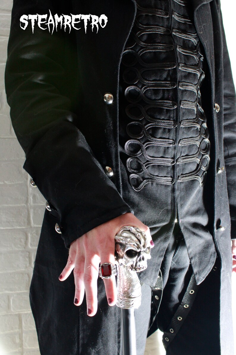 May include: A person wearing a black coat with a skull-shaped walking stick. The coat has a decorative design of black braid on the front. The person is wearing a ring with a red stone on their finger.