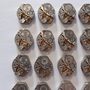 May include: A collection of 15 silver-toned watch movements with gold-colored gears. The watch movements are arranged in a grid pattern on a white surface.