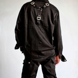 May include: Black long-sleeved shirt with a black harness and silver buckles. The shirt has a button-up collar and a single button on the front. The person is wearing black pants with multiple straps and buckles.