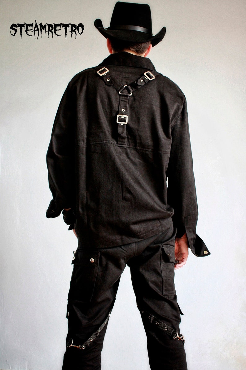 May include: Black long-sleeved shirt with a black harness and silver buckles. The shirt has a button-up collar and a single button on the front. The person is wearing black pants with multiple straps and buckles.