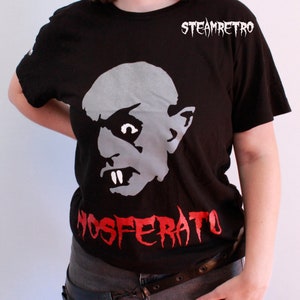 May include: Black t-shirt featuring a white graphic of a vampire from the film Nosferatu. The text "NOSFERATU" is printed in red below the graphic. The shirt has a crew neck and short sleeves.
