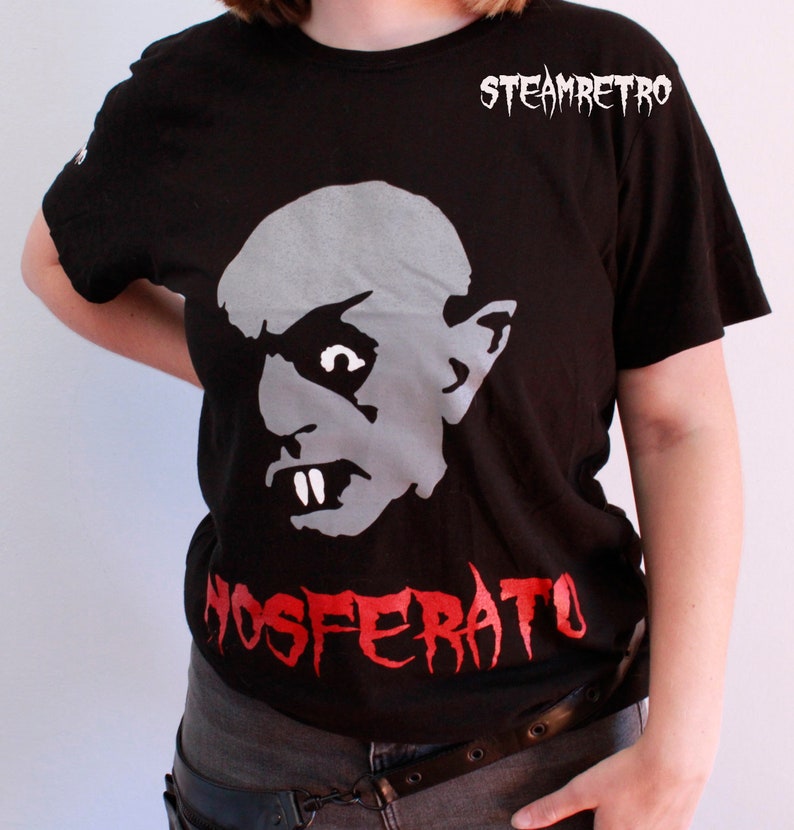 May include: Black t-shirt featuring a white graphic of a vampire from the film Nosferatu. The text "NOSFERATU" is printed in red below the graphic. The shirt has a crew neck and short sleeves.