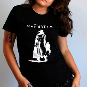 May include: A woman wearing a black t-shirt with a white graphic of a figure in a long coat and the text "Fields of the Nephilim". She is wearing black pants with silver buckles and rings on her fingers.