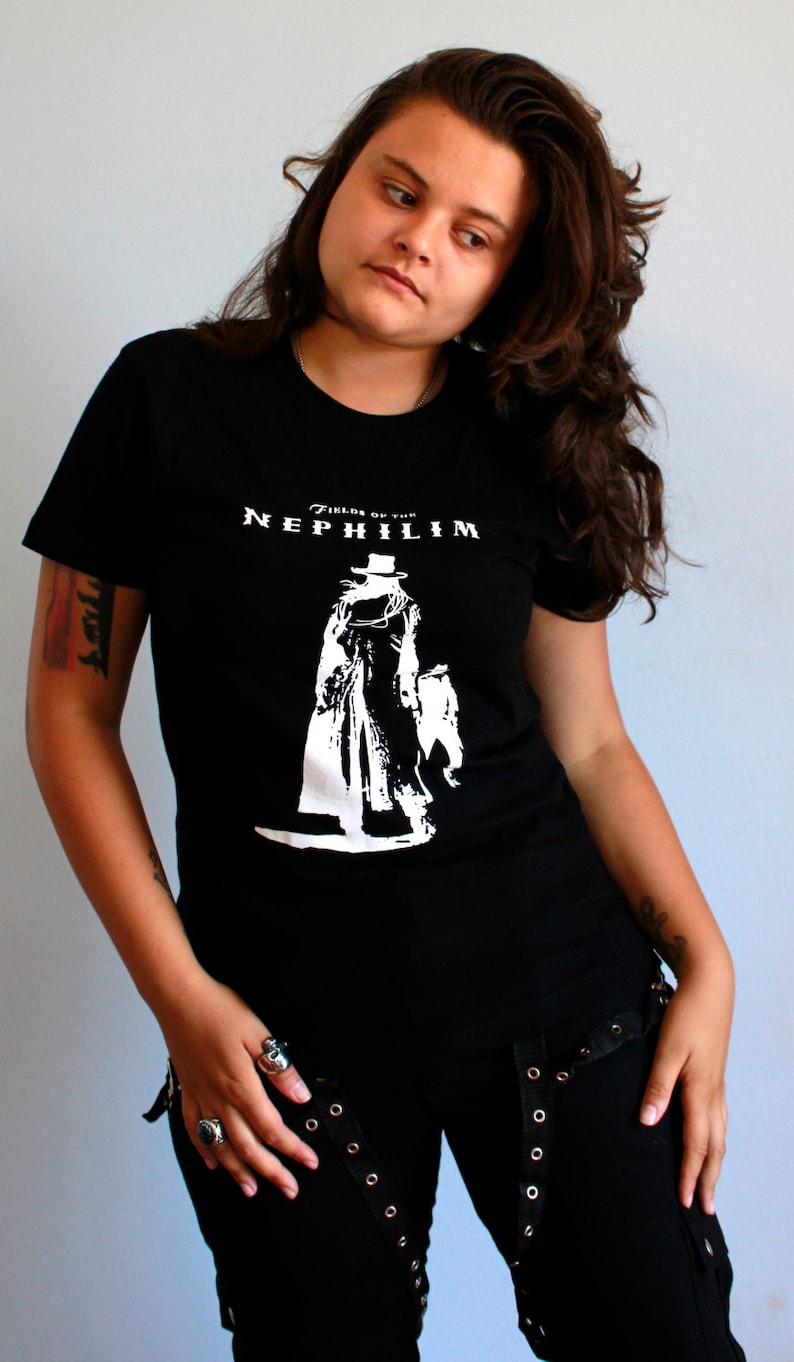 May include: A woman wearing a black t-shirt with a white graphic of a figure in a long coat and the text "Fields of the Nephilim". She is wearing black pants with silver buckles and rings on her fingers.