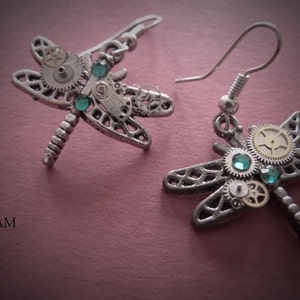 May include: A pair of silver dragonfly earrings with intricate clockwork details and green gemstones. The earrings are on a pink background.