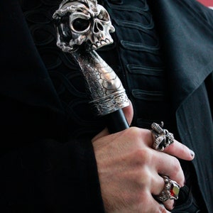 May include: A silver skull-shaped walking stick with a black handle. The stick is held by a hand wearing a silver skull ring and a red gemstone ring.