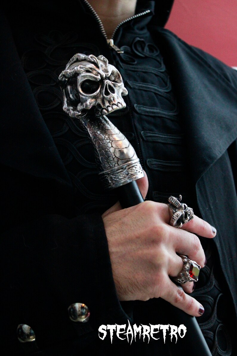 May include: A silver skull-shaped walking stick with a black handle. The stick is held by a hand wearing a silver skull ring and a red gemstone ring.