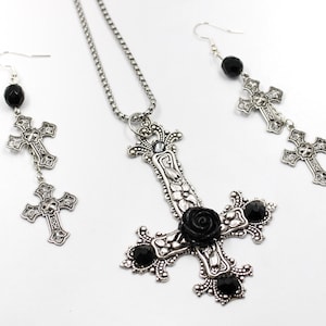 Puede incluir: Un conjunto de collar y pendientes de plata con un colgante de cruz invertida adornado con una rosa negra y detalles de piedras preciosas negras. Los pendientes tienen una perla negra y dos abalorios de cruz plateados.
