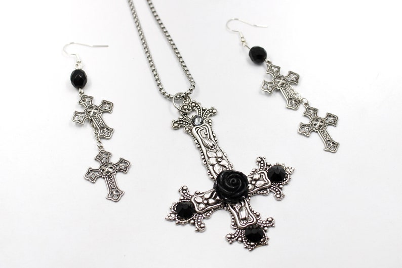 Puede incluir: Un conjunto de collar y pendientes de plata con un colgante de cruz invertida adornado con una rosa negra y detalles de piedras preciosas negras. Los pendientes tienen una perla negra y dos abalorios de cruz plateados.