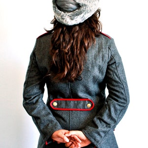 May include: A gray wool coat with a red trim and gold buttons. The coat has a long, fitted silhouette and a high collar. The model is wearing a gray fur hat with a black strap.
