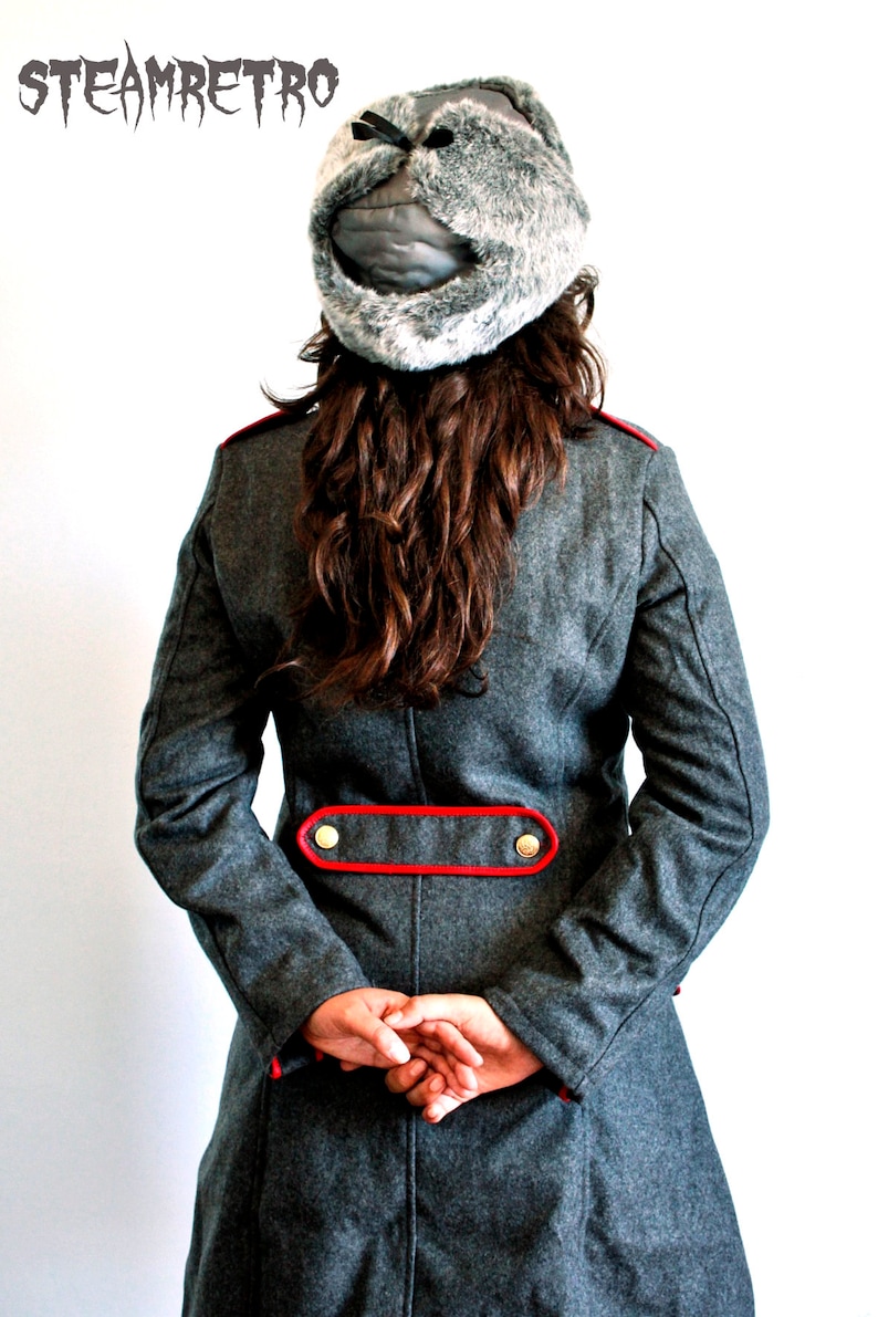 May include: A gray wool coat with a red trim and gold buttons. The coat has a long, fitted silhouette and a high collar. The model is wearing a gray fur hat with a black strap.