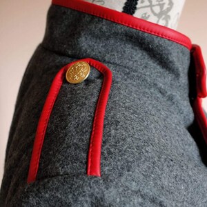 May include: Close-up of a gray wool jacket with red leather trim and a gold button. The jacket has a classic military style.