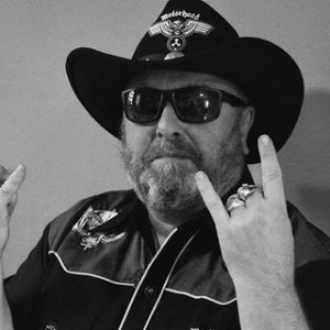 Könnte beinhalten: Schwarz-Weiß-Bild einer Person mit einem schwarzen Cowboyhut mit Motorhead-Logo, Sonnenbrille und Ringen. Die Person macht eine Rock'n'Roll-Handgeste. Die Person hat einen Bart und trägt ein schwarzes Hemd.