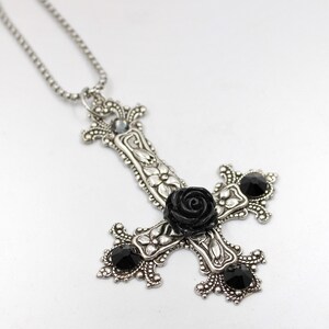 Puede incluir: Un collar con un colgante de plata en forma de cruz invertida adornado con una rosa negra y piedras preciosas negras.