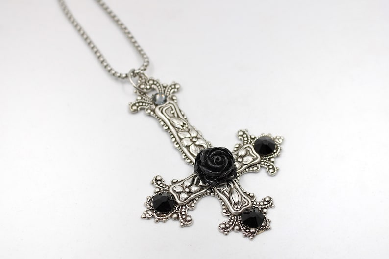 Puede incluir: Un collar con un colgante de plata en forma de cruz invertida adornado con una rosa negra y piedras preciosas negras.
